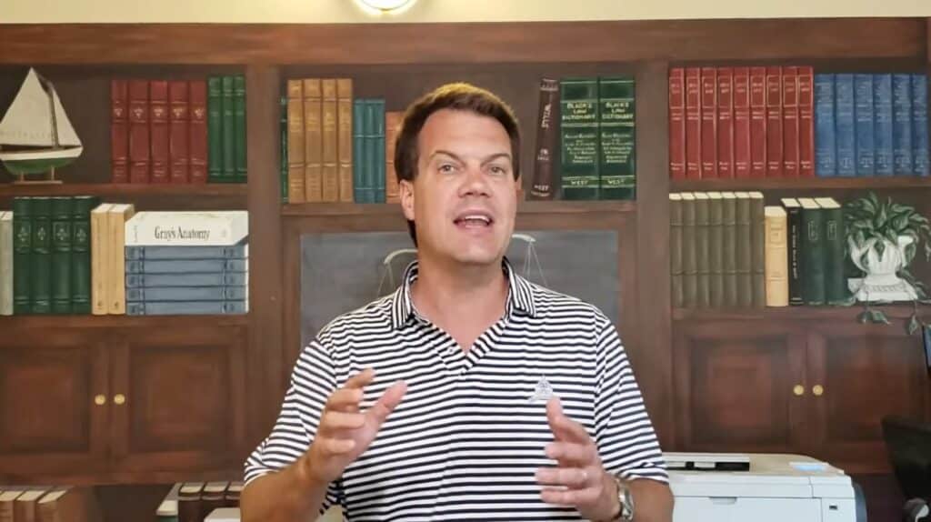 A man in a striped polo shirt speaks in front of a background designed to look like bookshelves and a sailboat model.
